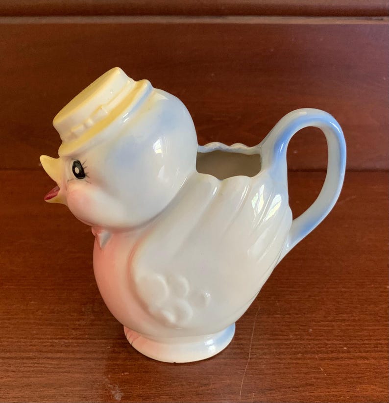 May include: A blue and pink ceramic creamer in the shape of a bird wearing a yellow hat. The creamer has a handle and a spout.
