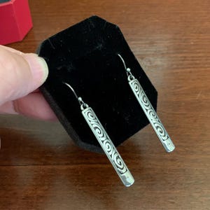 Puede incluir: Pendientes colgantes plateados con un dise&ntilde;o de barra rectangular. Cada pendiente presenta una serie de patrones en espiral grabados a lo largo de su longitud. Los pendientes se muestran sobre una superficie de terciopelo negro.