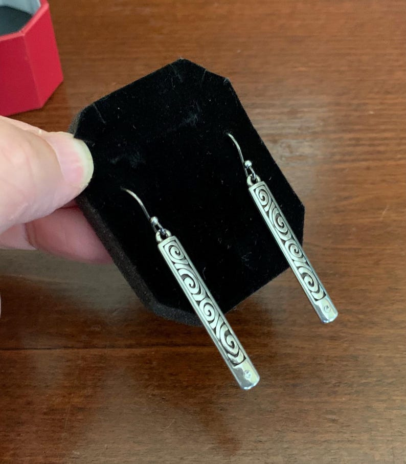 Puede incluir: Pendientes colgantes plateados con un dise&ntilde;o de barra rectangular. Cada pendiente presenta una serie de patrones en espiral grabados a lo largo de su longitud. Los pendientes se muestran sobre una superficie de terciopelo negro.