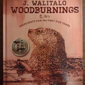 Op de afbeelding: Boekomslag voor "J. Walitalo Woodburnings" met de ondertitel "Highlights from the First Five Years". De illustratie toont een otter in het water, met een rond zegel "U.P. Notable Books". De naam van de auteur is Joanna Walitalo.