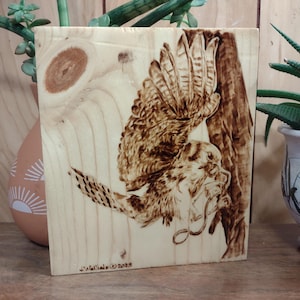 May include: A wooden plaque with a pyrography design of a bird in flight. The artwork is in shades of brown and tan, with the bird's wings outstretched. The artist's signature and the year 2023 are visible at the bottom.
