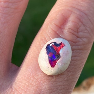 May include: A white oval-shaped opal cabochon with red, blue, and purple iridescent flashes. The opal is set in a white matrix.