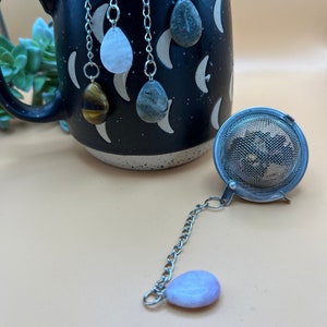May include: A set of four tea infusers with different colored gemstone charms. The charms are attached to silver chains and are hanging from the rim of a black mug with a moon pattern. A silver tea infuser with a mesh ball is also pictured.