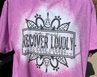 Puede incluir: Una camiseta morada con un diseño blanqueado. El diseño presenta un mandala con el texto "We must recover loudly to keep others from dying quietly."