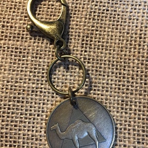 Op de afbeelding: Een goudkleurige sleutelhanger met een kameel silhouet in een driehoek. De sleutelhanger heeft een splitring en een karabijnhaak.