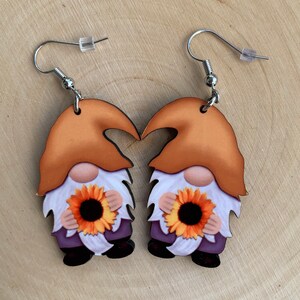 Peut inclure: Une paire de boucles d'oreilles en forme de gnome. Chaque boucle d'oreille représente un gnome avec un chapeau orange, une barbe blanche et un tournesol. Les gnomes portent des chemises violettes et des chaussures noires. Les boucles ont des crochets argentés.
