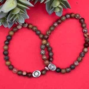 May include: Two brown and green beaded bracelets with silver charms. The charms are in the shape of a triangle with a circle inside. The bracelets are on a red background.