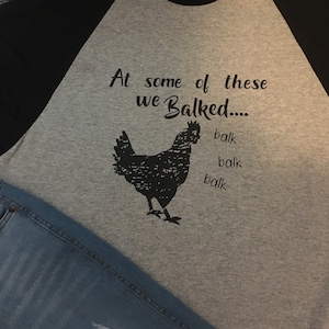 May include: A gray and black baseball tee shirt with the text "At some of these we Balked..." and a black silhouette of a chicken. The text "balk" is repeated three times below the chicken.