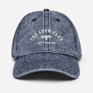 May include: A blue denim baseball cap with a white embroidered logo that reads "The Crow Club, Ketterdam".