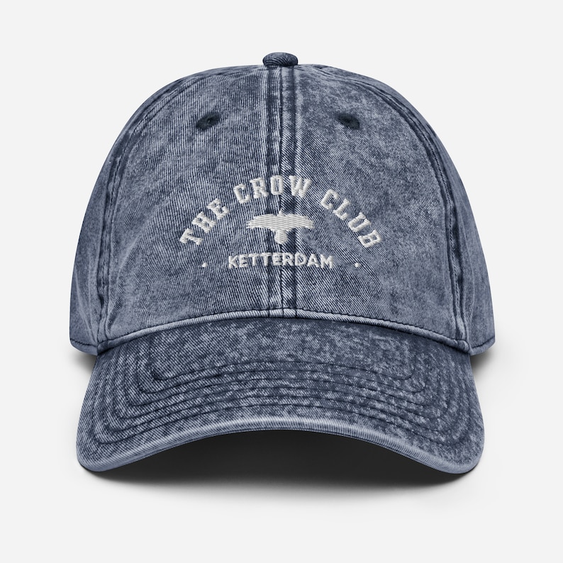May include: A blue denim baseball cap with a white embroidered logo that reads "The Crow Club, Ketterdam".