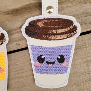 May include: A purple and white embroidered coffee cup keychain with a smiling face and brown lid.