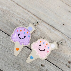 May include: Two ice cream cone-shaped keyrings with smiling faces. One is purple with orange and blue sprinkles, the other is pink with orange and blue sprinkles. Both have a white heart on the cone.