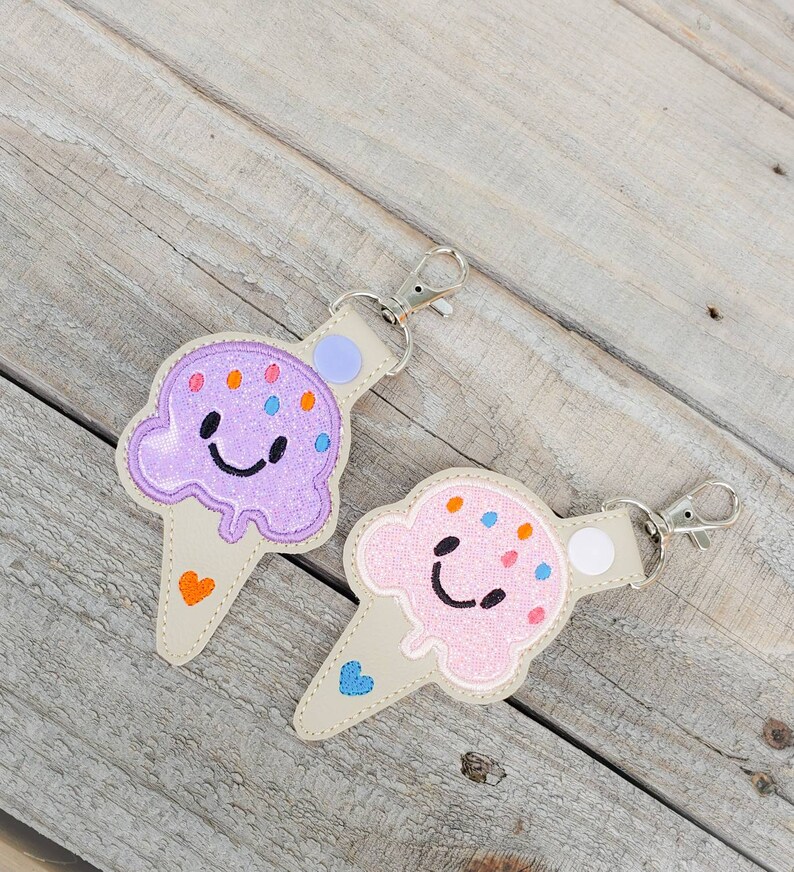 May include: Two ice cream cone-shaped keyrings with smiling faces. One is purple with orange and blue sprinkles, the other is pink with orange and blue sprinkles. Both have a white heart on the cone.