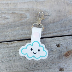 Peut inclure: Porte-clés en cuir blanc avec un nuage brodé bleu et blanc avec un visage souriant. Le porte-clés a un fermoir en métal argenté.