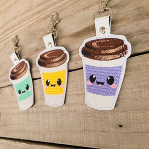 May include: Three embroidered coffee cup keychains with smiling faces. The cups are purple, yellow, and green with brown lids.