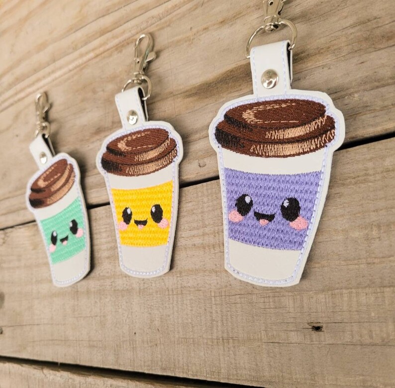 May include: Three embroidered coffee cup keychains with smiling faces. The cups are purple, yellow, and green with brown lids.