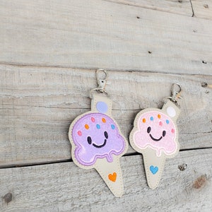 May include: Two ice cream cone keyrings with smiling faces. One is purple with orange and blue sprinkles and the other is pink with orange sprinkles. Both have a white heart on the bottom.