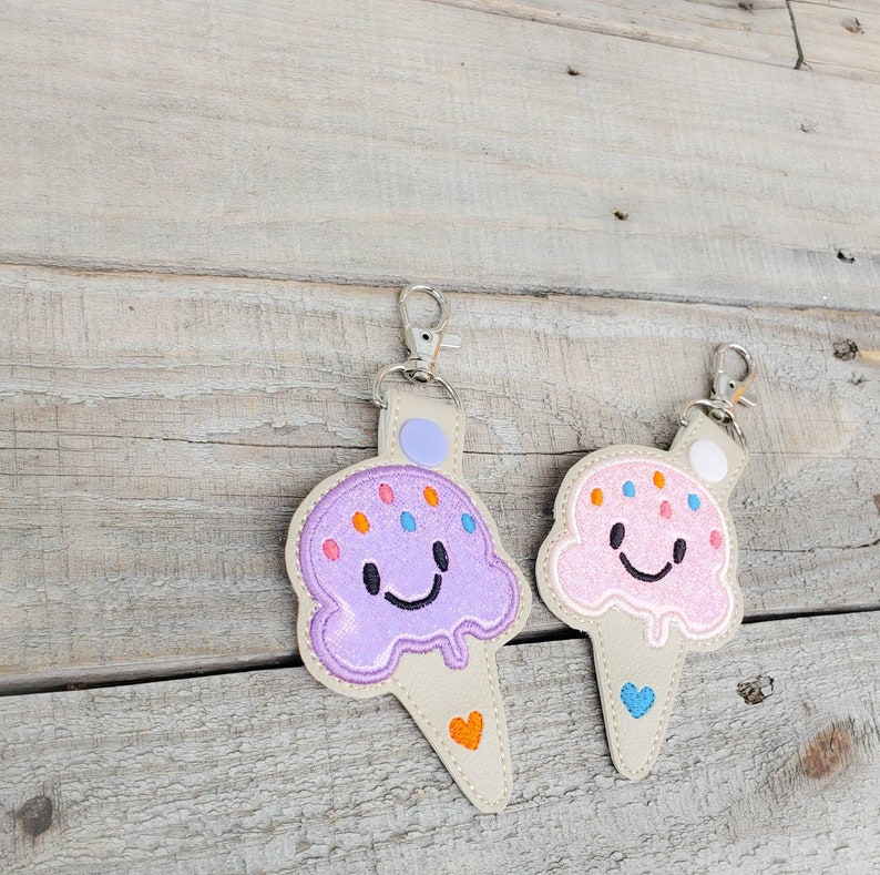 May include: Two ice cream cone keyrings with smiling faces. One is purple with orange and blue sprinkles and the other is pink with orange sprinkles. Both have a white heart on the bottom.