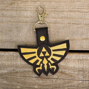 Peut inclure: Un porte-clés en cuir noir avec un symbole Triforce brodé en or. Le porte-clés a une boucle en métal argenté pour l'attacher aux clés.