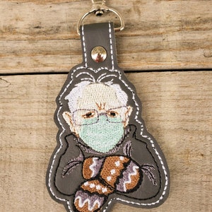 May include: A grey leather keychain featuring an embroidered image of a person wearing a face mask and holding mittens. The person has white hair and is wearing glasses.