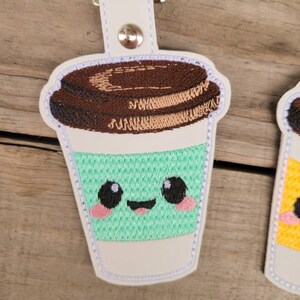 May include: A white, embroidered coffee cup keychain with a brown lid, green cup, and a cute cartoon face. The cup has a happy expression with black eyes, pink cheeks, and a red tongue.