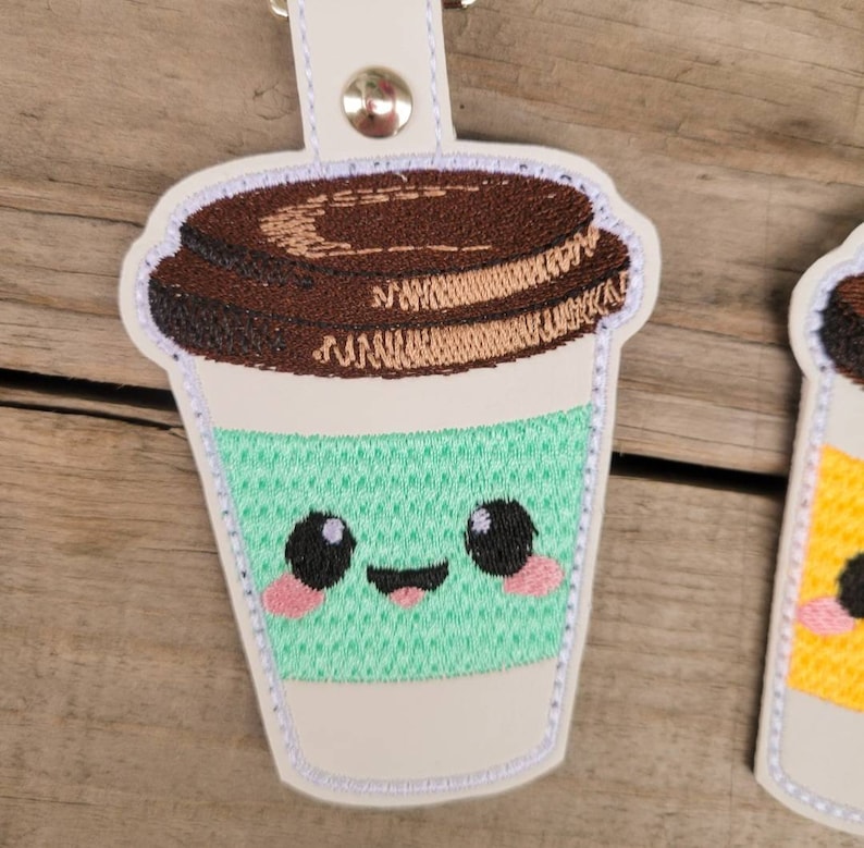 May include: A white, embroidered coffee cup keychain with a brown lid, green cup, and a cute cartoon face. The cup has a happy expression with black eyes, pink cheeks, and a red tongue.
