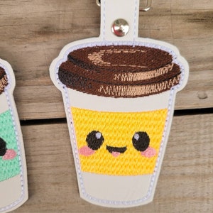 May include: A white, brown, and yellow embroidered coffee cup keychain with a smiling face. The cup has a brown lid and a yellow liquid inside.