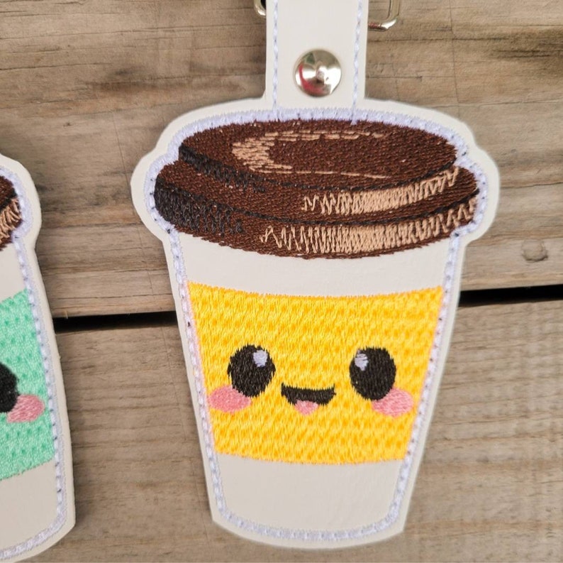 May include: A white, brown, and yellow embroidered coffee cup keychain with a smiling face. The cup has a brown lid and a yellow liquid inside.