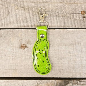 May include: A green glitter pickle keyring with a smiling face. The keyring has a silver metal clasp.