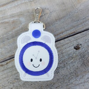 May include: White leather keychain with a blue button and an embroidered alarm clock design. The clock face has a smiling face with eyes and a mouth.