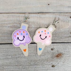 May include: Two embroidered ice cream cone keyrings with smiling faces. One is purple with orange sprinkles and the other is pink with blue sprinkles.