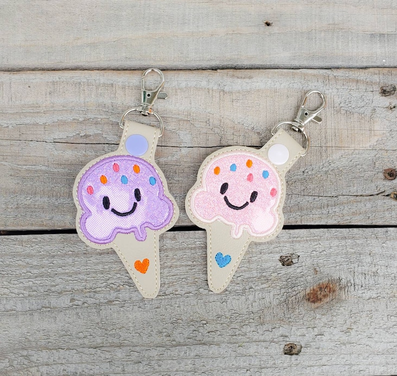 May include: Two embroidered ice cream cone keyrings with smiling faces. One is purple with orange sprinkles and the other is pink with blue sprinkles.