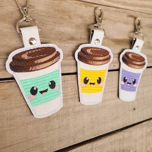 May include: Three embroidered coffee cup keychains with smiling faces. The cups are green, yellow, and purple. Each keychain has a brown lid and a silver metal ring.