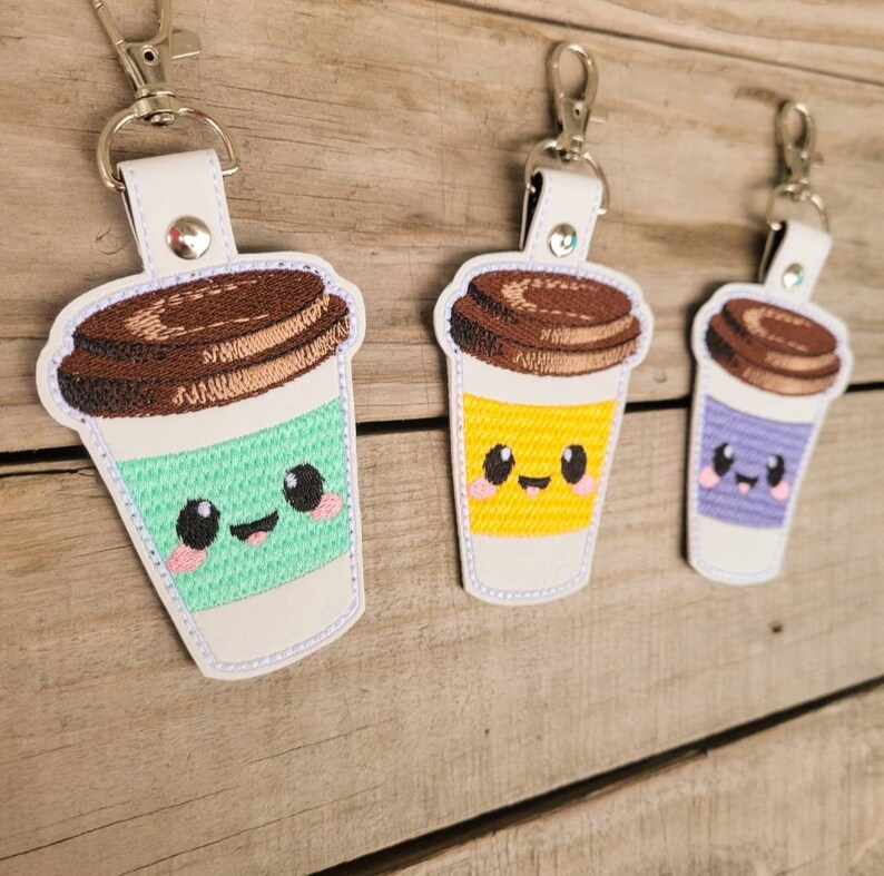 May include: Three embroidered coffee cup keychains with smiling faces. The cups are green, yellow, and purple. Each keychain has a brown lid and a silver metal ring.