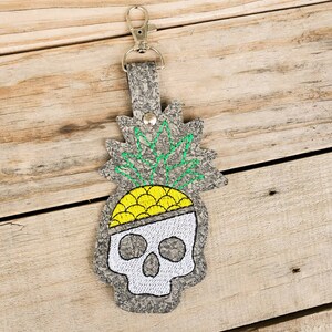 May include: A gray leather keychain with a skull design. The skull is topped with a yellow pineapple with green leaves.