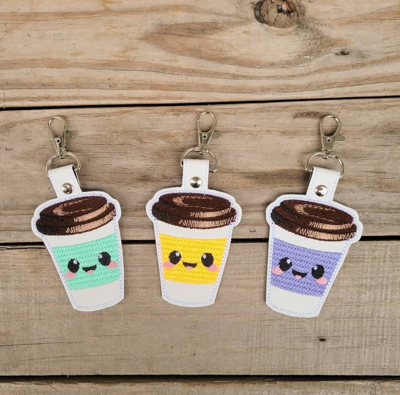 May include: Three embroidered coffee cup keychains with cute faces. The coffee cups are green, yellow, and purple. Each keychain has a silver metal ring.