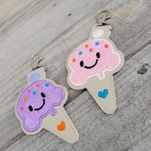 May include: Two ice cream cone-shaped keychains with smiling faces. One is pink with blue sprinkles and a blue heart, the other is purple with orange sprinkles and an orange heart.