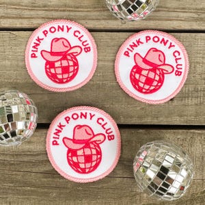 May include: Three pink and white embroidered iron-on patches with the text "Pink Pony Club" and a pink disco ball with a pink cowboy hat on top.