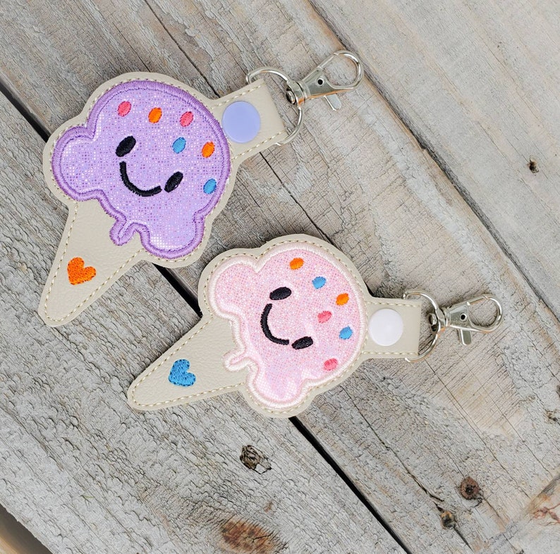 May include: Two ice cream cone-shaped keyrings with smiling faces. One is purple with orange and blue sprinkles, the other is pink with orange and blue sprinkles. Both have a white button and a silver keyring.