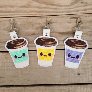 May include: Three embroidered keychains shaped like coffee cups with smiling faces. The cups are green, yellow, and purple. Each keychain has a brown lid and a silver metal ring.