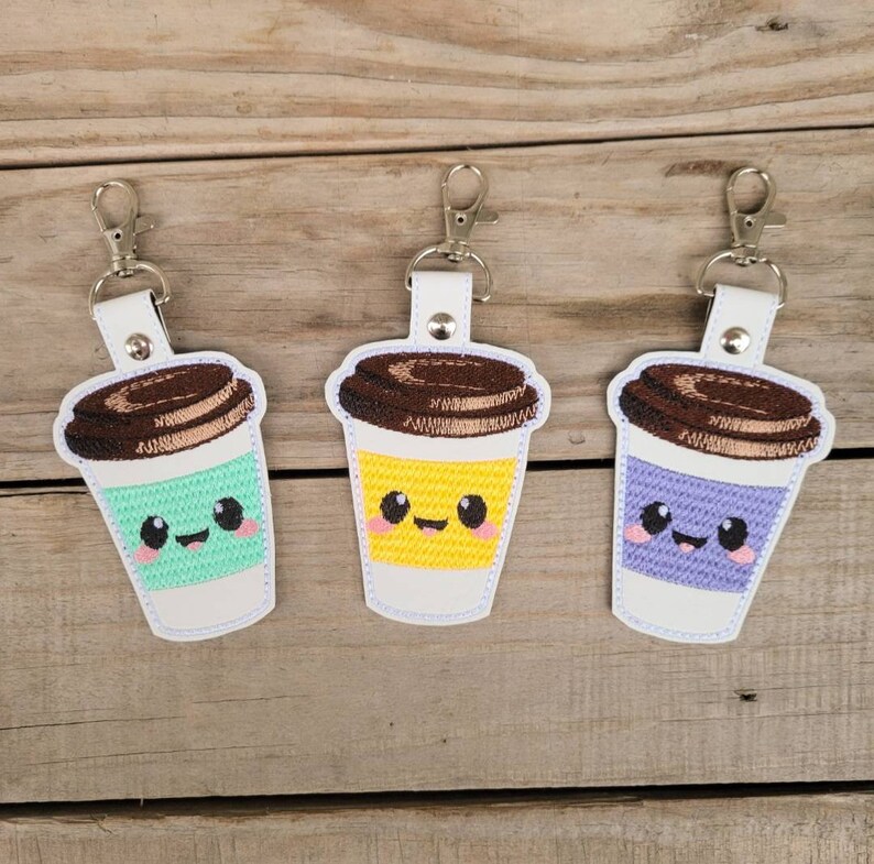 May include: Three embroidered keychains shaped like coffee cups with smiling faces. The cups are green, yellow, and purple. Each keychain has a brown lid and a silver metal ring.