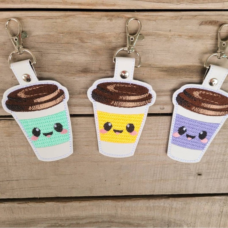 May include: Three embroidered coffee cup keychains with cute faces. The coffee cups are white with brown lids and different colored liquid inside. The keychains are attached to silver key rings.