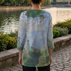 May include: A long-sleeved jacket featuring a print of a woman with a green umbrella, inspired by a classic painting. The jacket's design includes a serene landscape with figures, in shades of blue, green, and white. The jacket is worn outdoors.