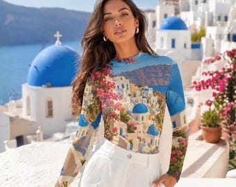 May include: A long-sleeved top featuring a scenic print of a coastal village with white buildings and blue domes. The top has a crew neck and is paired with white pants. The image is set against a backdrop of a blue sea and sky, with pink flowers.
