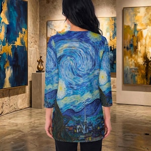 May include: A long-sleeved tunic featuring a print of Van Gogh's "Starry Night" painting. The tunic has a blue and yellow color palette, with a swirling night sky design. The bottom of the tunic depicts a village with a church.