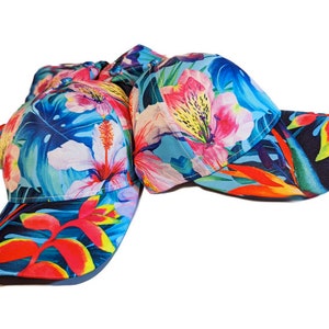 Op de afbeelding: Twee baseballpetten met een tropische bloemenprint. De petten zijn blauw, groen, roze, oranje en geel. De print bevat hibiscusbloemen, palmbladeren en andere tropische planten.