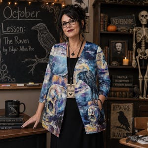 May include: A long-sleeved open-front blazer featuring a vibrant, painterly print of ravens and human skulls in shades of blue, purple, and yellow. The garment has a straight hem, three-quarter length sleeves, and side pockets, worn over a solid black dress.