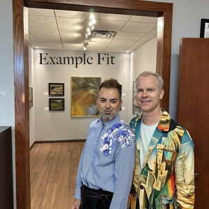 May include: A light blue pinstriped button-down shirt with blue and white floral embroidery on the left shoulder, paired with black trousers. Beside it, a blazer featuring a colorful surrealist landscape print over a light blue t-shirt and dark pants.