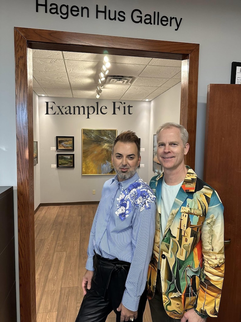 May include: A light blue pinstriped button-down shirt with blue and white floral embroidery on the left shoulder, paired with black trousers. Beside it, a blazer featuring a colorful surrealist landscape print over a light blue t-shirt and dark pants.