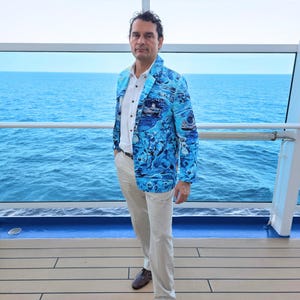 May include: A bright blue blazer featuring a detailed nautical print with small boats and waves, worn over a crisp white button-down shirt and light beige trousers. The blazer has a notched lapel, long sleeves, and a structured fit with dark buttons.